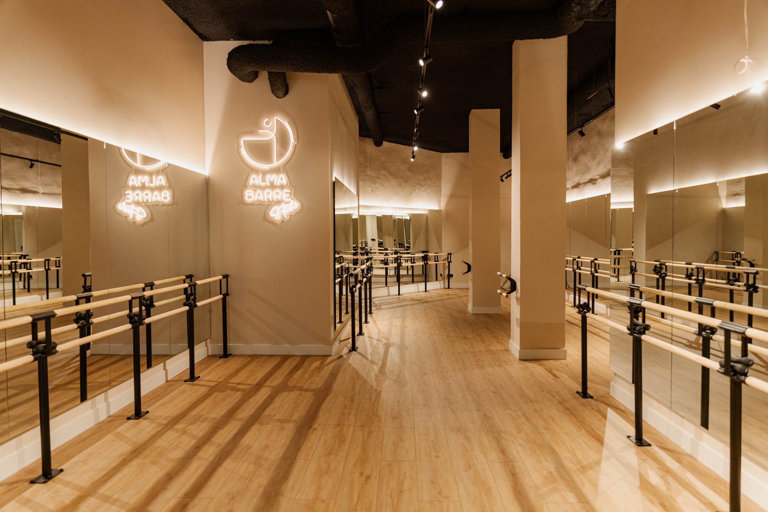 Sala de barre del estudio boutique con el logo de Alma Barre iluminado, con barras dobles, suelos de madera y espejos